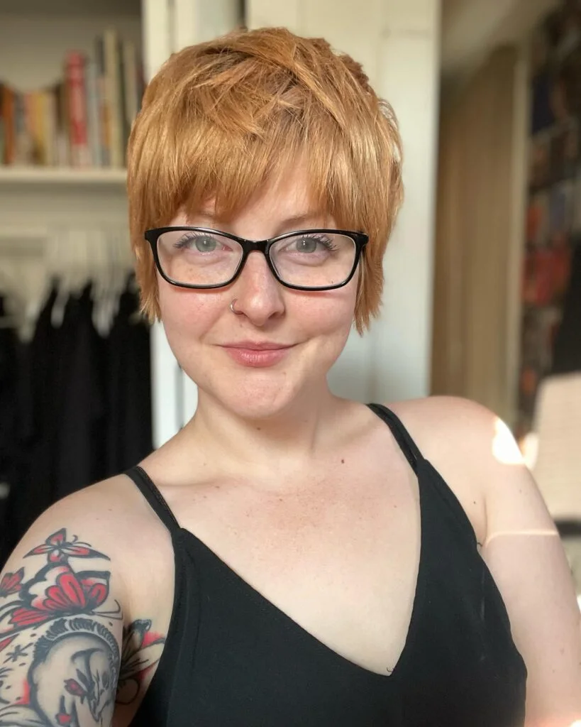 straight copper pixie with a lifted top, close sides, and light fringe above black glasses on a round face with thin hair