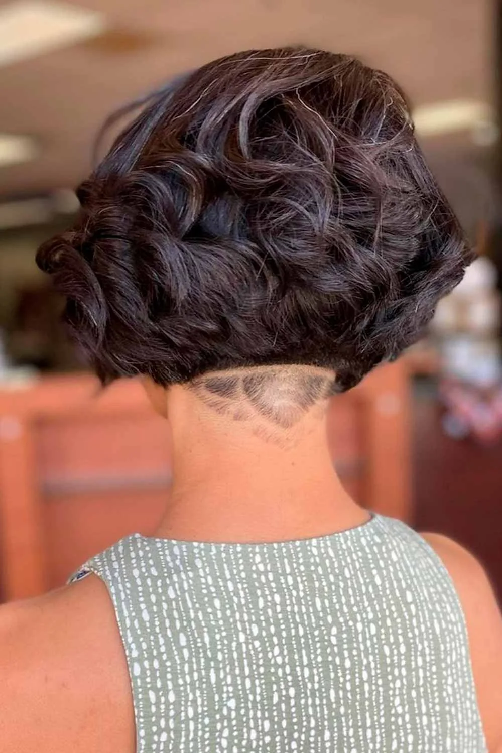 Back view of a short dark curled bob with a shaved nape design
